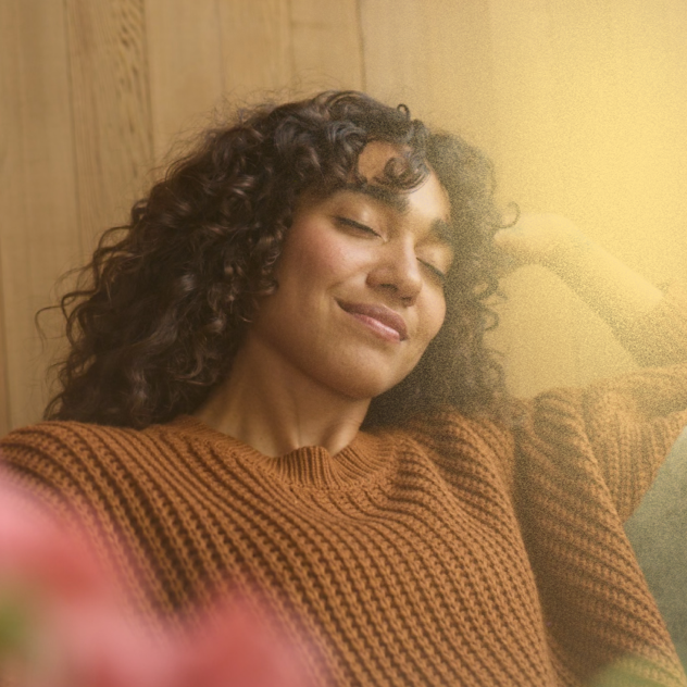 Woman with curly hair smiling, eyes closed, wearing a rust-orange sweater in warm light.