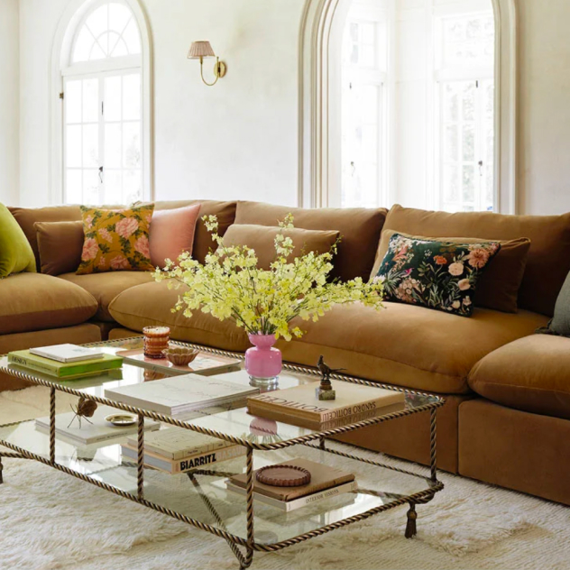 Camel velvet sofa with floral pillows, a glass coffee table with books, and a vase of yellow flowers.