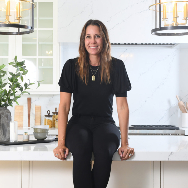 Becki Owens smiling in a white marble kitchen with brass pendant lighting.