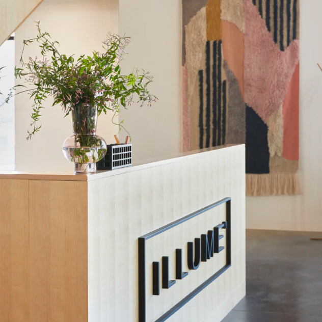 Reception desk with a 3D 'ILLUME' sign, a vase of greenery, and a woven wall hanging.