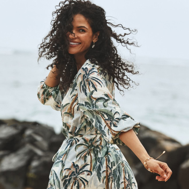Woman with curly hair smiling in a palm-print dress on a rocky beach.