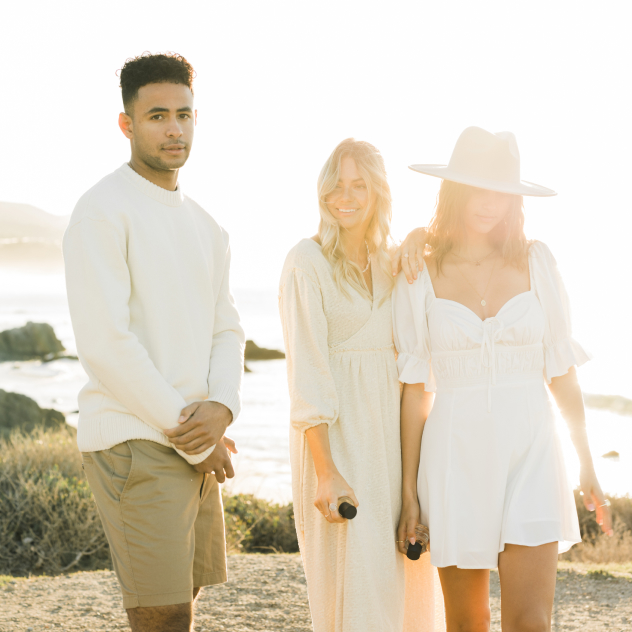 Three people in neutral, cream-colored clothing standing together on a sunlit coastal bluff.