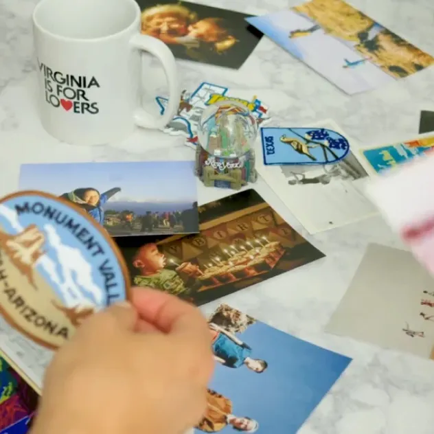Hand holding a travel patch over a table scattered with keepsakes, postcards, and personal photos.