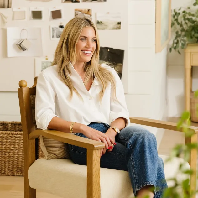 Studio McGee co-founder Shea McGee smiling, seated in a wooden armchair in a bright, styled living room.