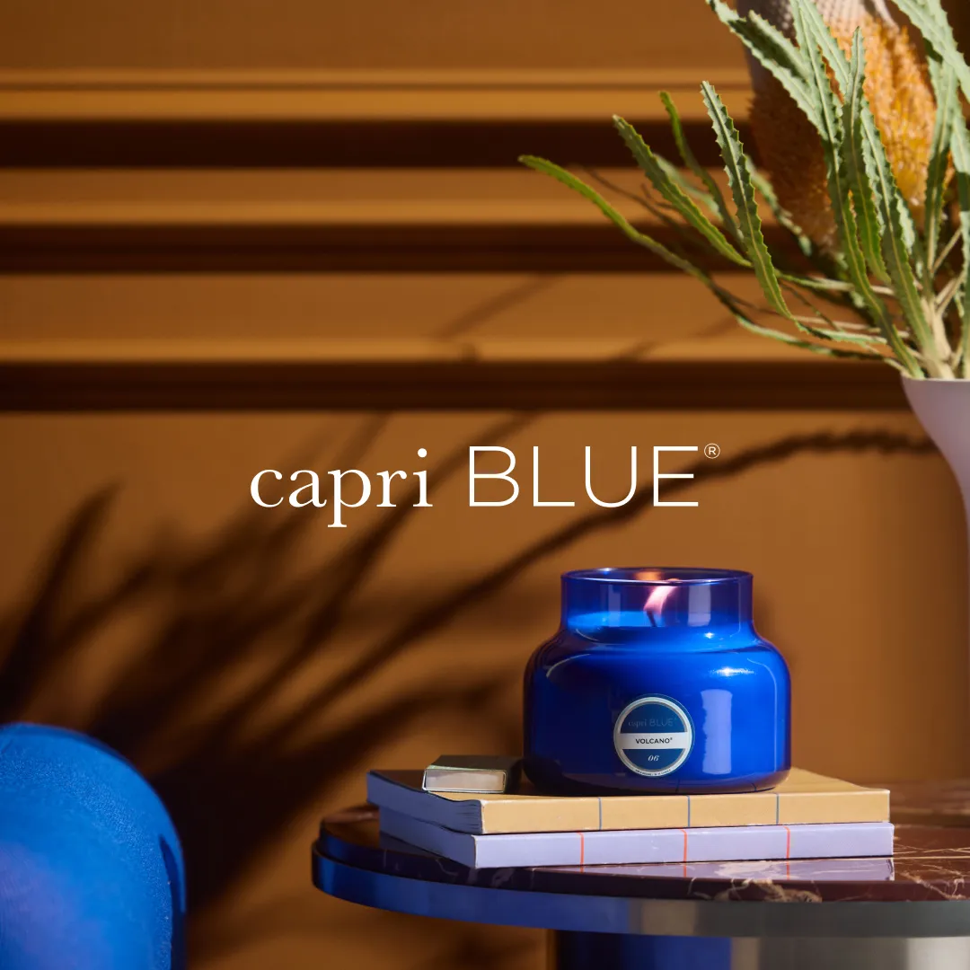 Capri Blue Volcano candle on stacked books, with the 'capri BLUE' wordmark overlaid on the wall behind it.