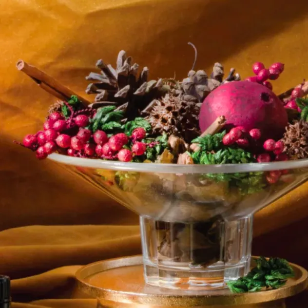 Glass compote filled with pinecones, cinnamon sticks, holly berries, and a pomegranate on gold fabric.