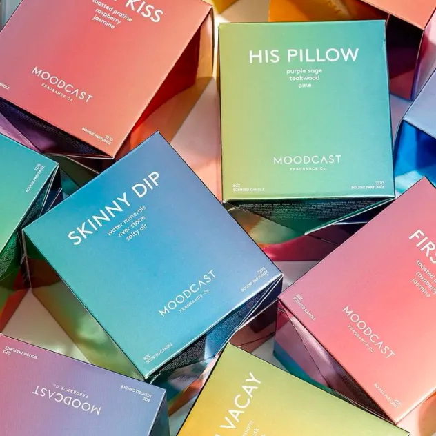 Pile of colorful iridescent Moodcast fragrance boxes, including 'Skinny Dip' and 'Vacay'.