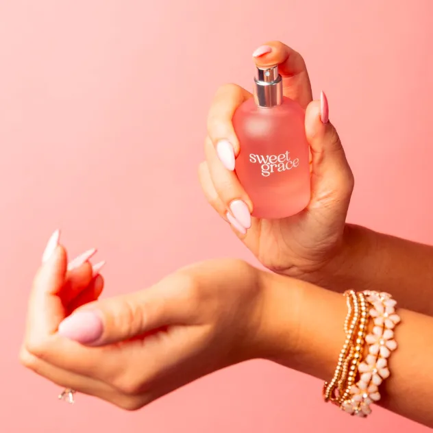 Hand holding a pink 'sweet grace' perfume bottle above another open hand, against a pink background